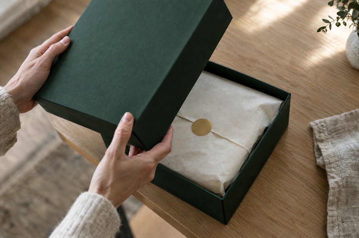 Hands lifting the lid off a green box to reveal wrapped tissue paper